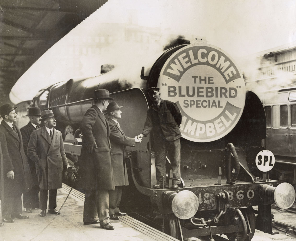 Sir Malcolm Campbell shakes hands with the driver of a steam engine with a special plaque on the front reading, 'Welcome Campbell - The Bluebird Special'. Also in the picture, far left, is Cuthbert Grasemann, PR and Publicity Officer for Southern Railways. Presumably he is the one responsible for this meeting. Date: c.1927
