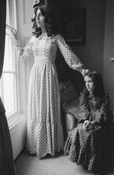 Fashion -- a woman and a girl, both wearing full-length Laura Ashley style dresses in floral fabric, looking out of a window. Date: circa 1970s