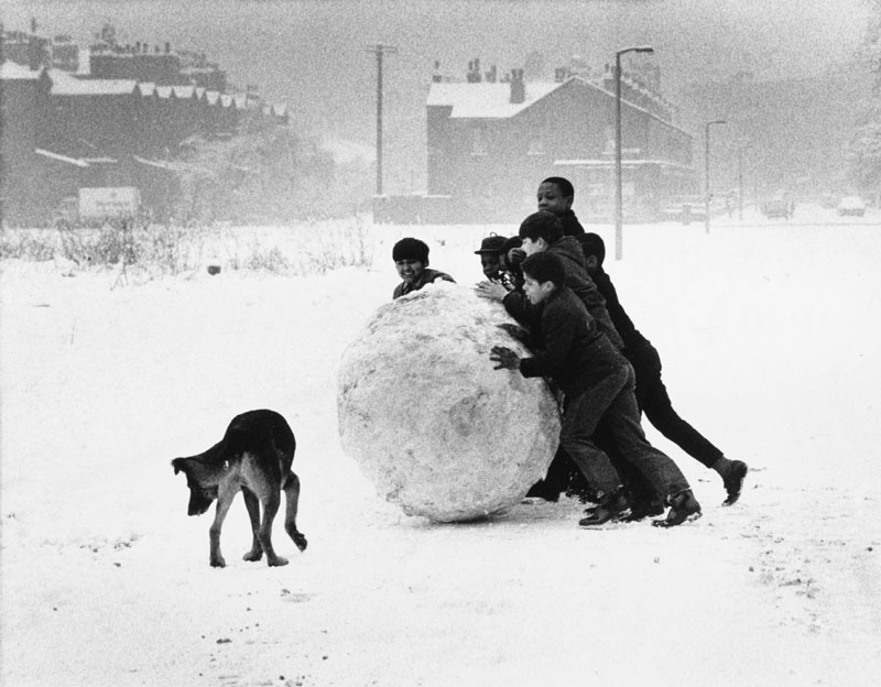 A group of black and Asian boys make a massive snowball on a wintry Manchester day. Photograph by Shirley Baker Date: 1968