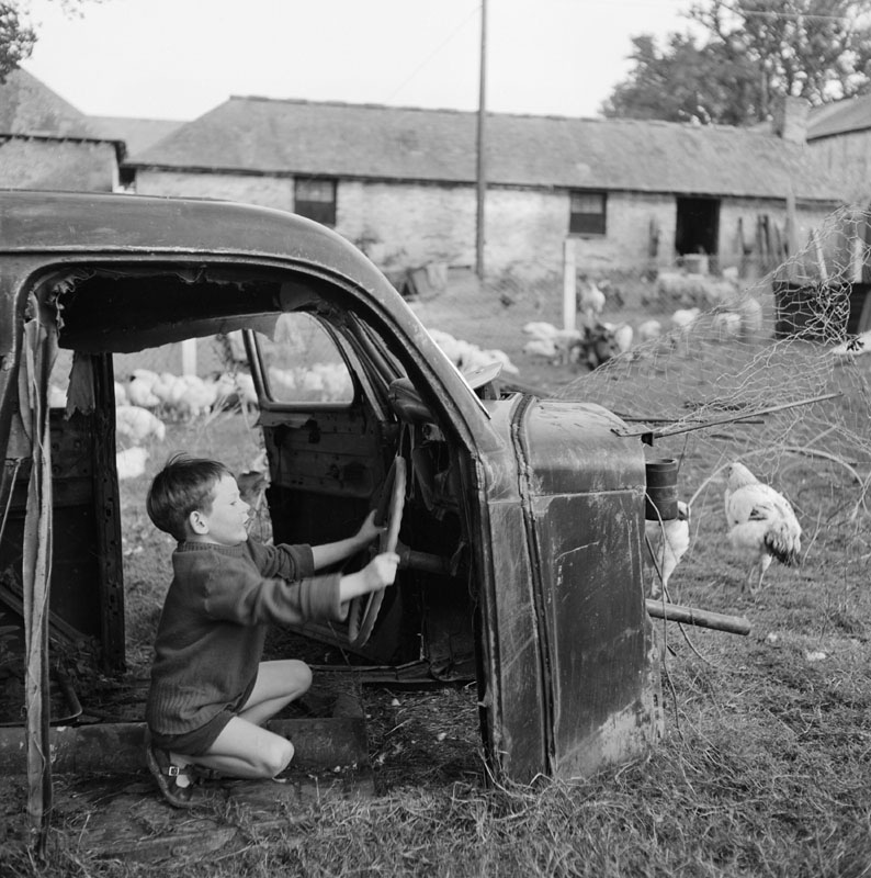 A young boy plays in the shell of an old car in a farmyard Date: 01 Jan 1955 - 30 Apr 1957