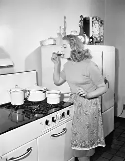 Sipping: Young woman in an apron in her kitchen tasting her